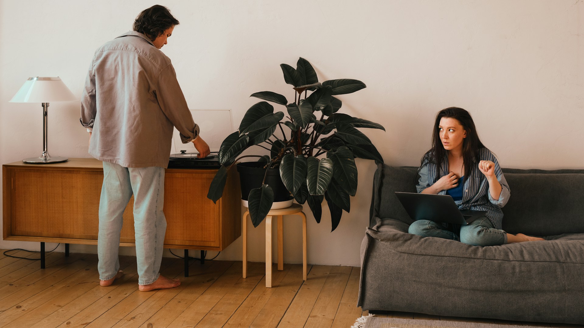 Couple in a home setting, man adjusting a record player while woman sits on a couch with a laptop, reflecting domestic dynamics.